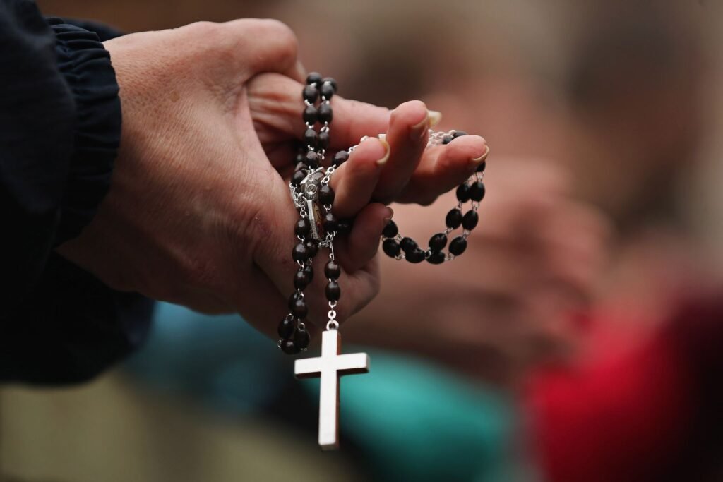 A close-up of hands tightly holding a traditional dark-beaded Christian rosary with a silver cross, serving as a symbolic visual for how addressing Christian persecution forms Trump’s new foreign policy roadmap.
