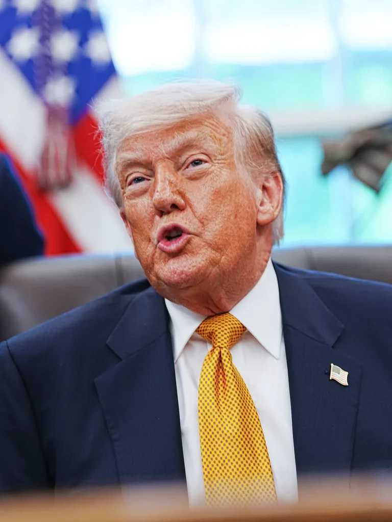 A close-up portrait of Donald Trump speaking emphatically, featuring the American flag in the blurred background, used to discuss the administration's stance on an Iran Naval Blockade and the resulting global pressure.