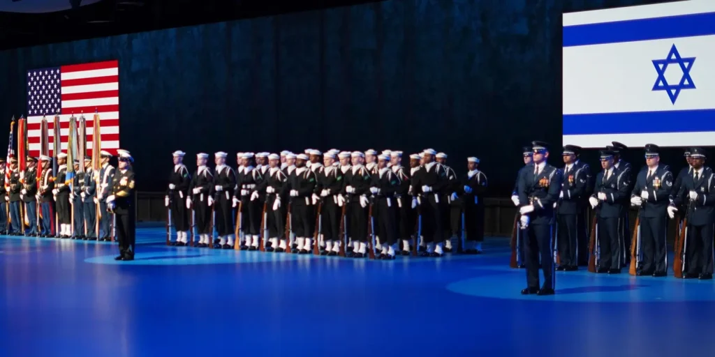 A large formation of military honor guard members standing at attention on a blue-lit stage flanked by a massive United States flag on the left and an Israeli flag on the right, providing visual context for how Congress quietly moves to integrate US-Israeli militaries.