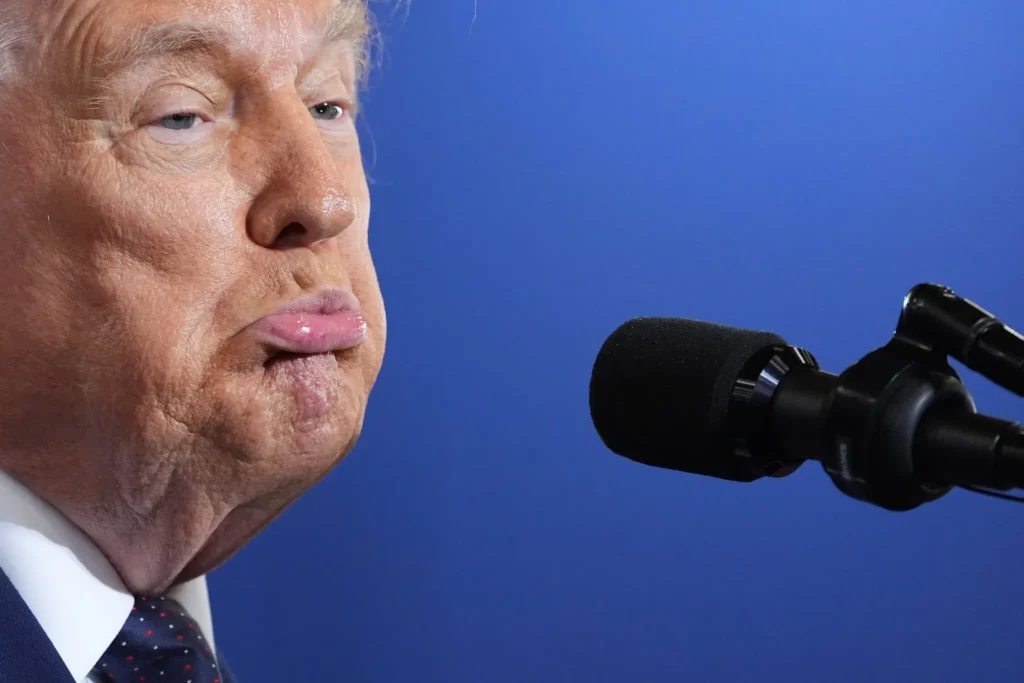 A tight close-up profile shot of Donald Trump with a tight expression speaking into a black microphone against a solid blue background, visually capturing the tense political moments described in It's Crow.
