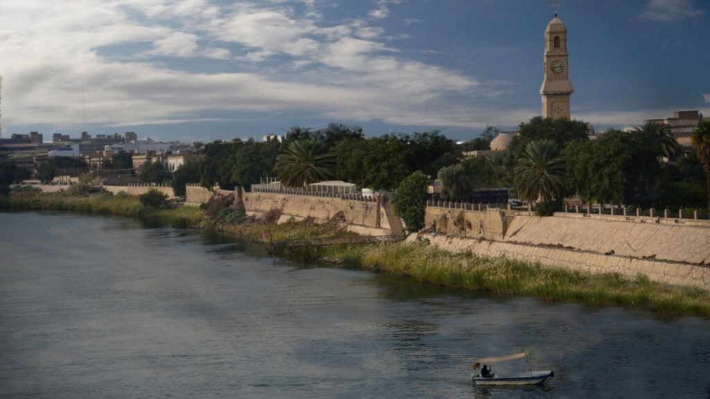 Scenic view of a historic Iraqi riverfront with a clock tower and traditional boat, illustrating the cultural backdrop against which Iraq corruption Ali al-Zaidi fails the nation and undermines national heritage.