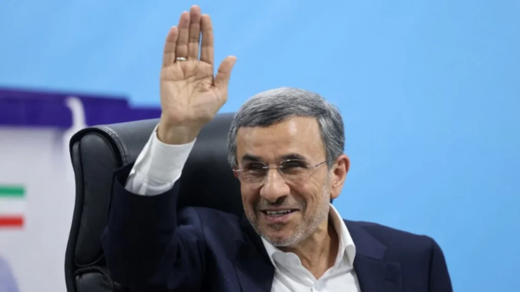 Former Iranian President Mahmoud Ahmadinejad smiling and waving his hand while seated in a black leather office chair against a solid light blue background, providing context for the geopolitical debates surrounding the alleged Iran threat.