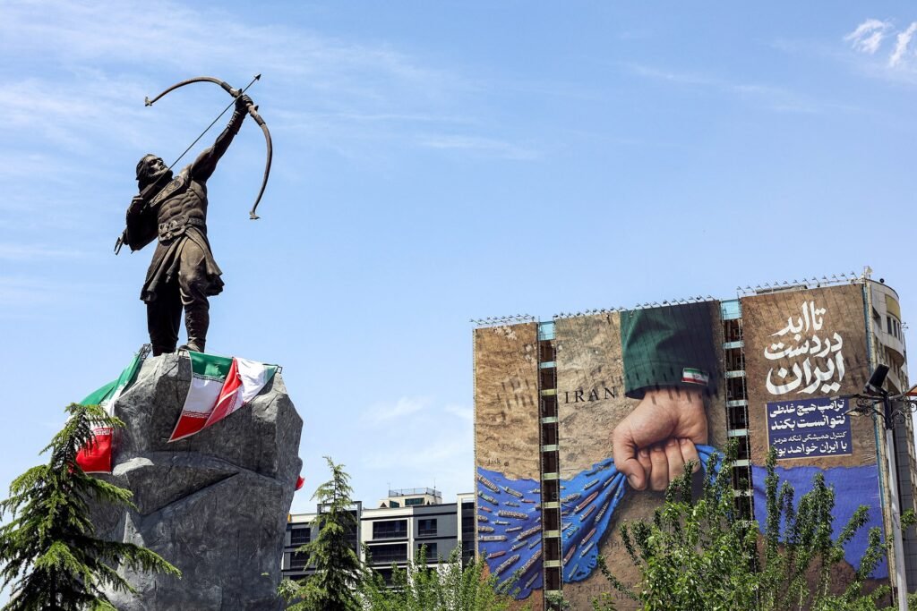 The bronze statue of Arash the Archer draped in Iranian flags stands prominently in Tehran, positioned next to a large geopolitical mural depicting a fist controlling the Strait of Hormuz, symbolizing historical and modern Iran war lessons for global strategy.