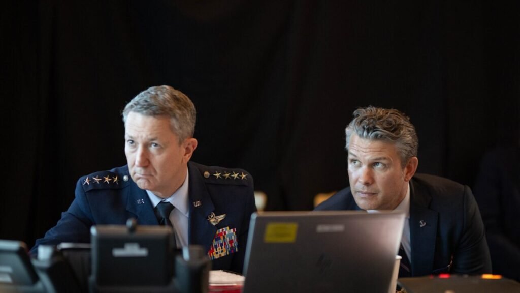 U.S. Air Force General Dan Caine in full dress uniform and Secretary of Defense Pete Hegseth sit behind laptops during a strategic operational briefing, focusing on operational metrics that intersect with concerns over civilian casualties.