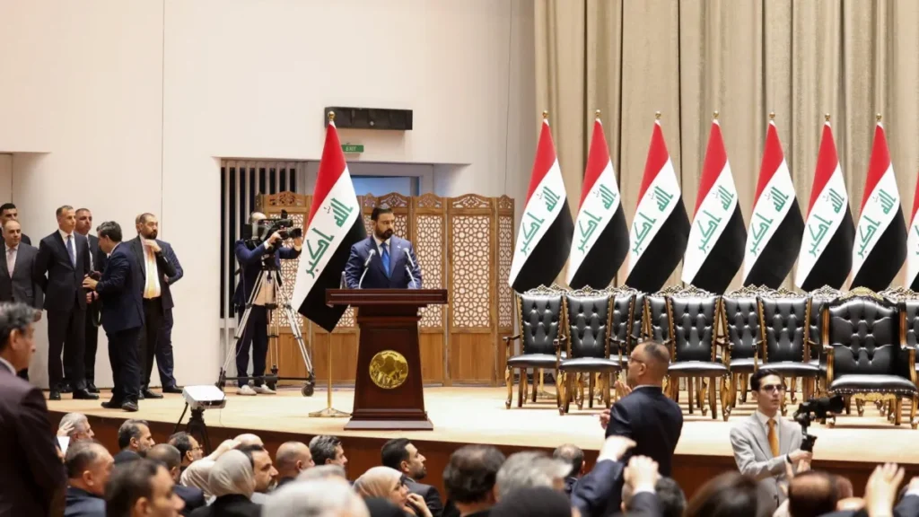 Iraq’s New Premier Begins Tenure Without Iran’s Allies A wider perspective of the Iraqi parliamentary hall showing Prime Minister-designate Ali al-Zaidi at the podium, surrounded by Iraqi flags and seated officials, capturing the institutional setting of the Iraq government deadlock.