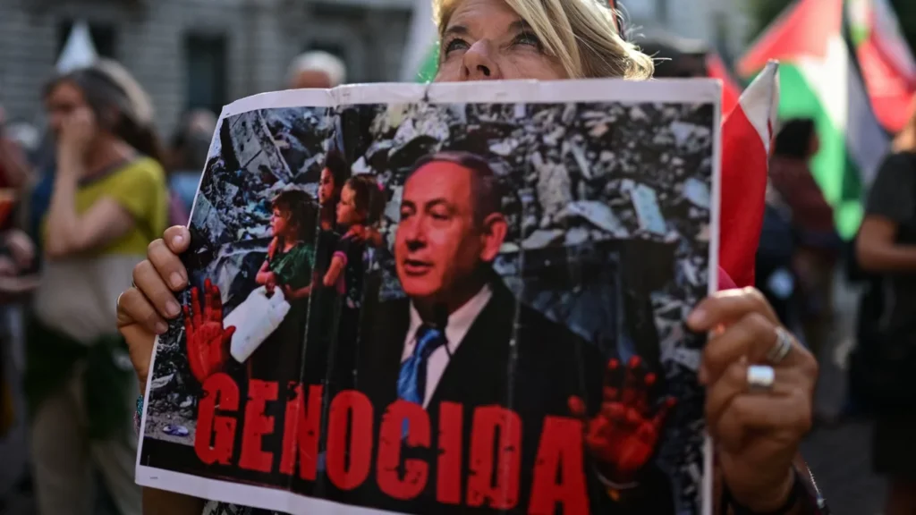 A demonstrator holds a protest sign featuring a portrait of Benjamin Netanyahu superimposed over background ruins with the text "GENOCIDA", highlighting the severe international backlash surrounding the prime minister's regional wartime visit.