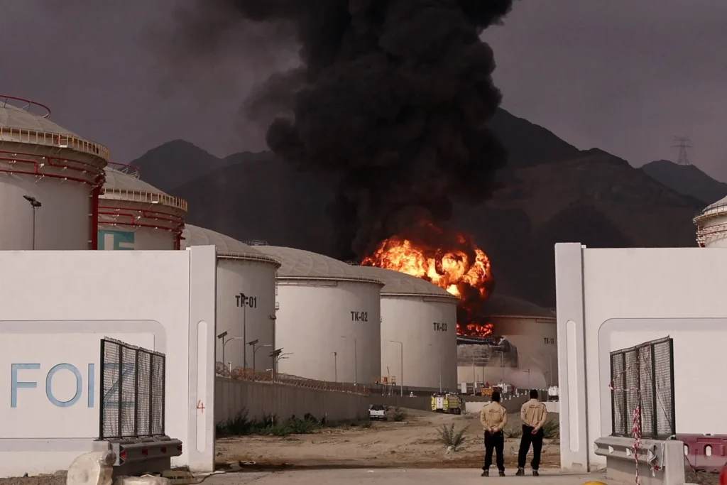 The Iran problem won’t be solved without a counter-drone coalition Security personnel stand near the entrance of an industrial energy storage facility with white fuel tanks labeled TK-01 and TK-02, as a massive explosion and thick black smoke erupt from a facility tank targeted in a regional strike, illustrating the security crises driving the need for a counter-drone coalition.