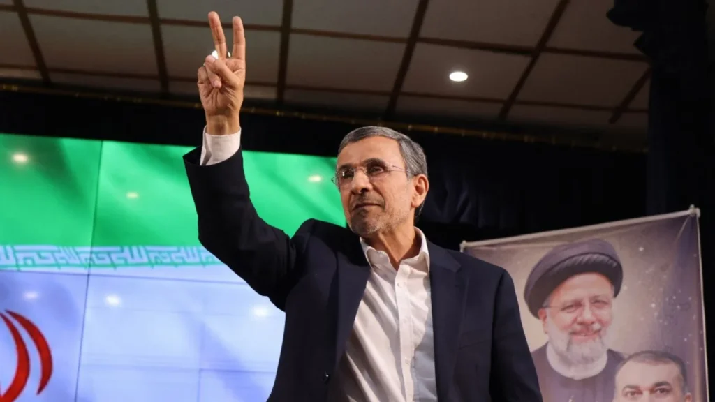 A political photo illustration depicting Mahmoud Ahmadinejad raising a peace sign in front of a green backdrop, juxtaposed with media narratives that analyze modern geopolitical theater using tropes like the 'Baby Shah'.