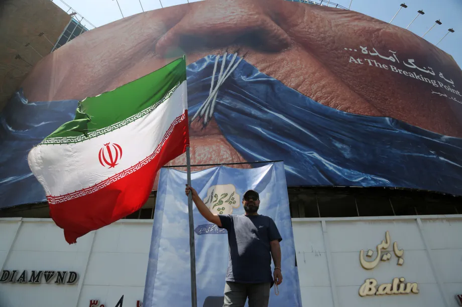 إيران، ترامب، والشقوق في القوة الأمريكية A man stands holding a large Iranian flag beneath a massive urban billboard depicting a symbolic maritime choke point, illustrating local resilience and geopolitical resistance amid shifting dynamics of American power.