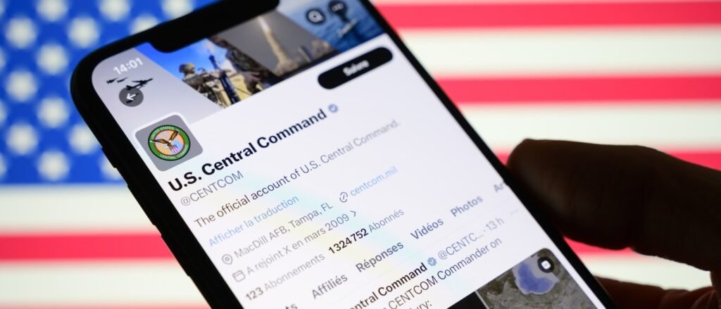 A hand holding a smartphone displaying the official U.S. Central Command (CENTCOM) X (formerly Twitter) profile against the backdrop of an American flag, symbolizing the front lines of the Iran Information War and the digital battle for narrative control.