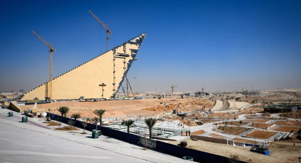 A sweeping view of a vast development site in Riyadh with a giant triangular landmark under construction and active yellow cranes, illustrating the infrastructure scale and execution challenges of Saudi Arabia Vision 2030.