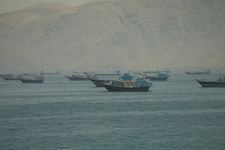 How the end of the nuclear file could reshape Iran, and the region A maritime view of various cargo vessels and dhows anchored in the Strait of Hormuz against the hazy backdrop of the mountainous Musandam coastline in Oman, symbolizing the regional economic channels tied to the future of the nuclear file.