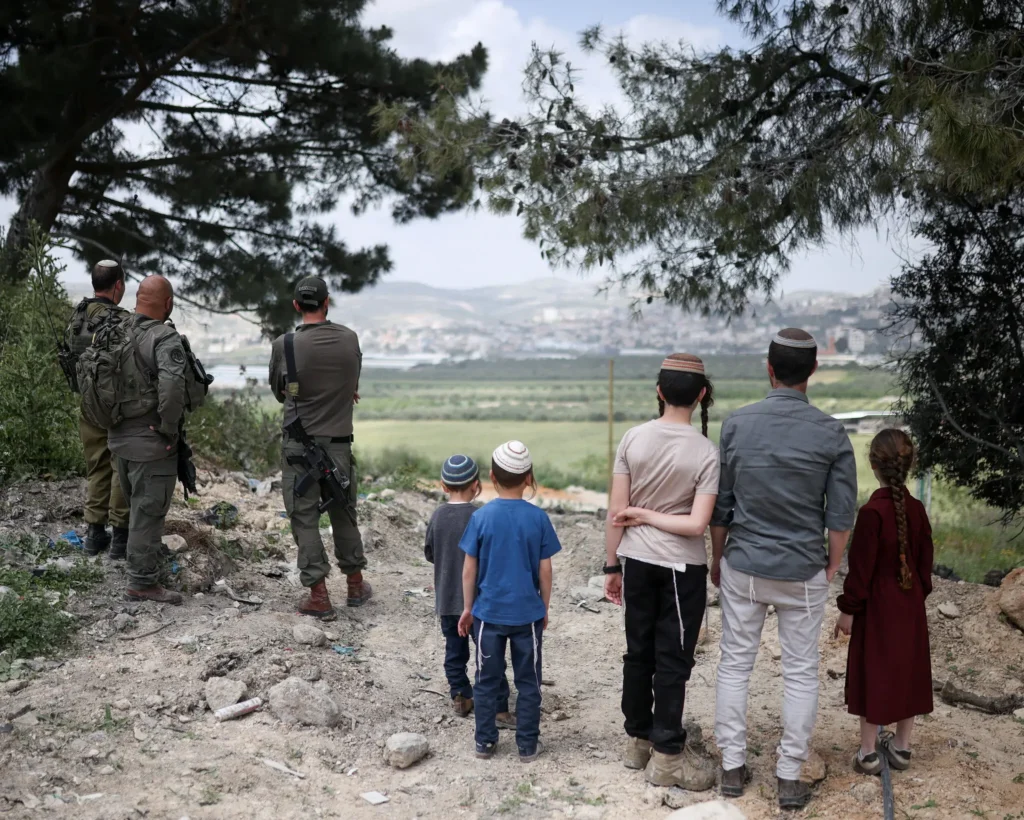 A group of families and children look out over a valley toward a distant town, standing near security personnel on a rocky hillside, representing the regional hope for stability under the Trump Plan to Save Lebanon.