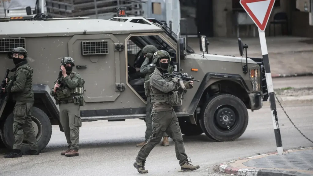 آیا سیانان بهتازگی آذربایجان را بهعنوان شریک نظامی مخفی اسرائیل افشا کرده است؟ Several armed Israeli soldiers in full green tactical gear and balaclavas patrol a street next to a large military armored vehicle, providing visual context to investigations regarding covert operations and the regional footprint of a secret military partnership.