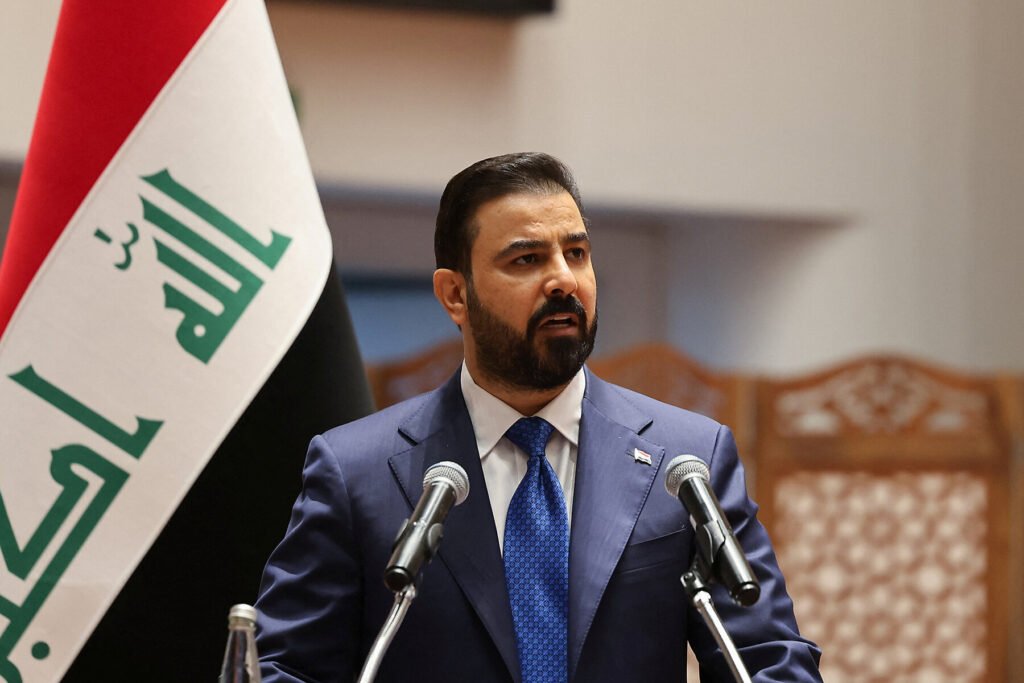 A portrait of Iraq's new Prime Minister Ali Al Zaidi wearing a blue suit and silk tie while speaking at a podium with microphones, with the national flag of Iraq on the left, capturing the diplomatic outreach central to repairing Iraq-Gulf relations.