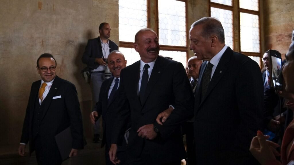 Azerbaijani President Ilham Aliyev in a dark suit smiling while walking arm-in-arm with Turkish President Recep Tayyip Erdogan at an international summit, surrounded by diplomats and officials inside a hall with large windows, illustrating the shifting diplomatic dynamics between Turkey and Azerbaijan.