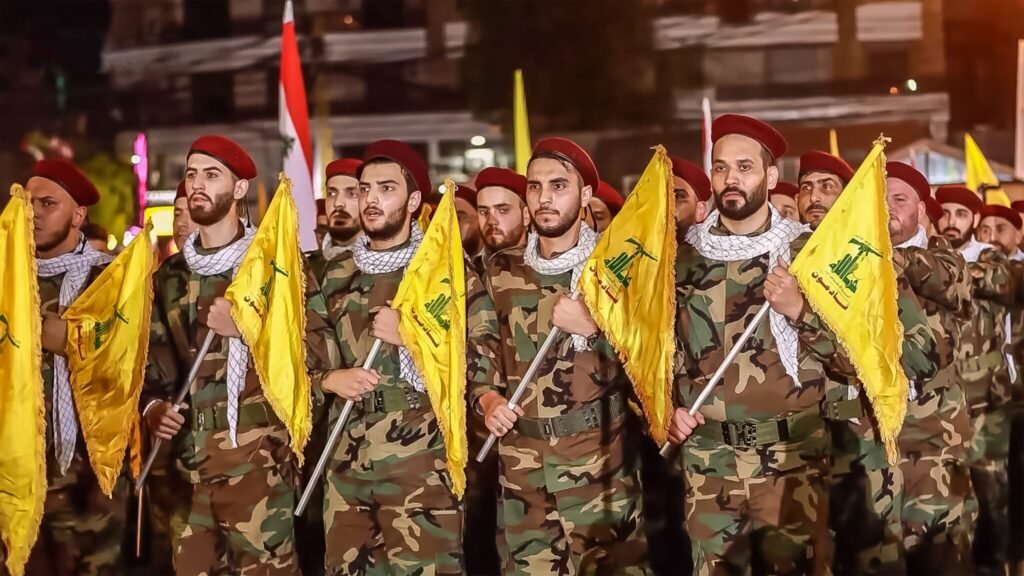 A row of Hezbollah members in camouflage uniforms, red berets, and traditional kaffiyehs standing in formation while carrying yellow flags during a public gathering, highlighting the military presence intertwined with diplomatic outcomes like An Iran Deal.