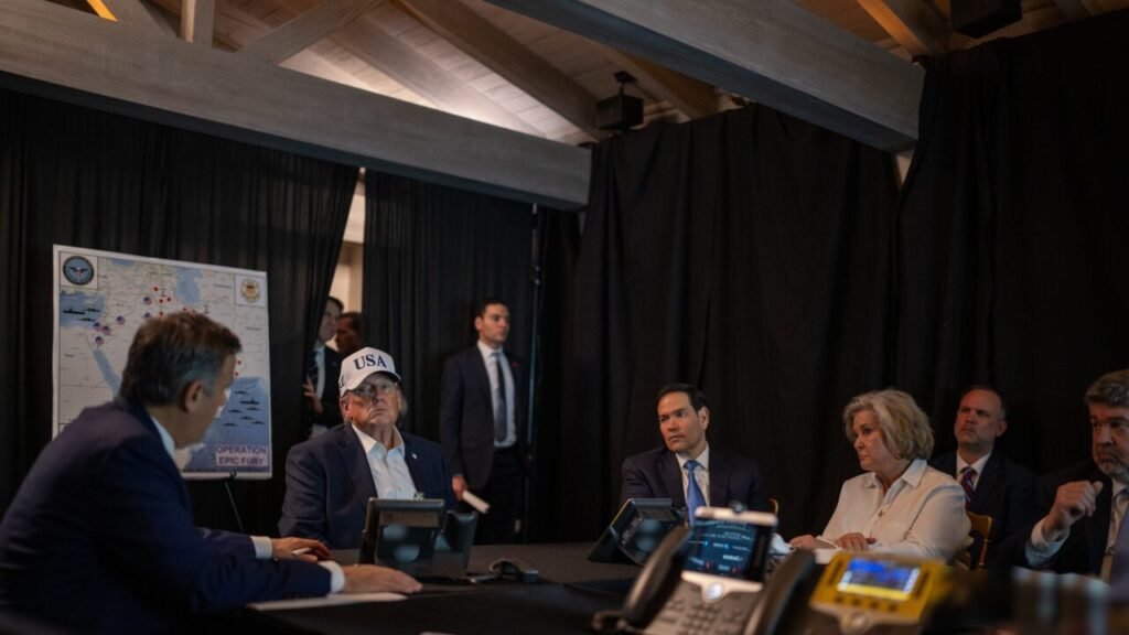 US President Donald Trump wearing a white USA cap looks toward an official during a briefing next to an "Operation Epic Fury" map display, with Marco Rubio and other advisors seated at a conference table, illustrating strategic recalculations after The US Lost the Iran War.