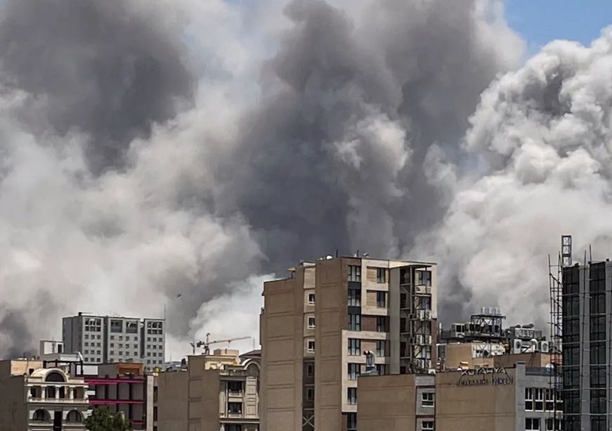 Russia’s Strategic Calculus After the Strikes on Iran Thick plumes of gray and white smoke rising high into the sky behind a skyline of multi-story residential buildings in Tehran, captured after an explosion to illustrate the regional security shifts impacting Russia Iran relations.