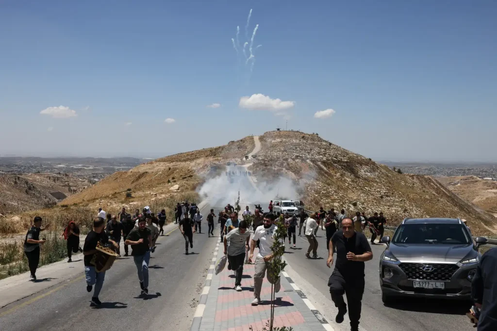The cycle of violence: From Israel-Palestine to Iran and back A large crowd of people fleeing down a paved hill road next to a silver SUV, with thick white smoke filling the path behind them and white smoke trails in the clear blue sky above, capturing a tense moment in the ongoing cycle of violence.
