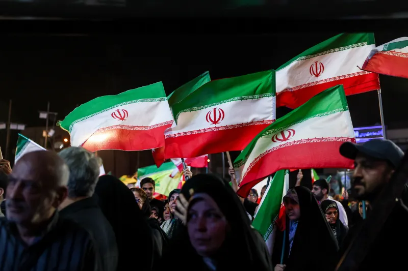 Iran Embraces a Forever War A night view of a crowd in Tehran waving multiple large Iranian flags during a public demonstration, capturing the ideological and nationalist mobilization reflecting how Iran Embraces a Forever War.