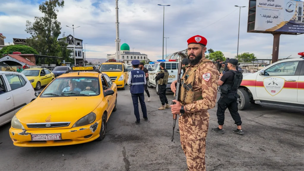 An Army of “Sons of the Regions”: Syria’s Post-Assad Military Order A Syrian military police officer with a beard and a red beret holding a rifle on a busy city street in Damascus, flanked by yellow taxis, traffic police officers, and white patrol vehicles, capturing the local security presence establishing Syria's post Assad military order.