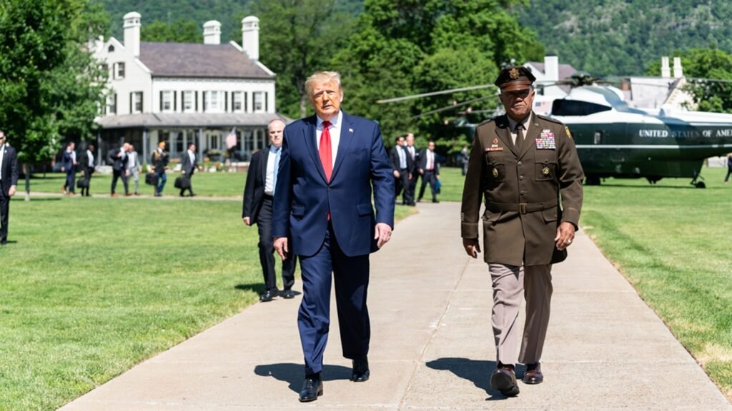 U.S. President Donald Trump in a blue suit and red tie walking along a path next to a decorated military officer in a brown uniform at West Point, with Marine One and official staff in the background, illustrating the strategic posturing evaluated against Iran's Mafia State.