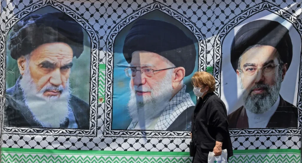 Washington and Tehran’s Very Dangerous Moment A masked pedestrian walks past a massive street banner displaying portraits of Ayatollah Ruhollah Khomeini, Ayatollah Ali Khamenei, and Mojtaba Khamenei in Tehran, visually grounding the geopolitical tension behind Washington and Tehran's Dangerous Moment.
