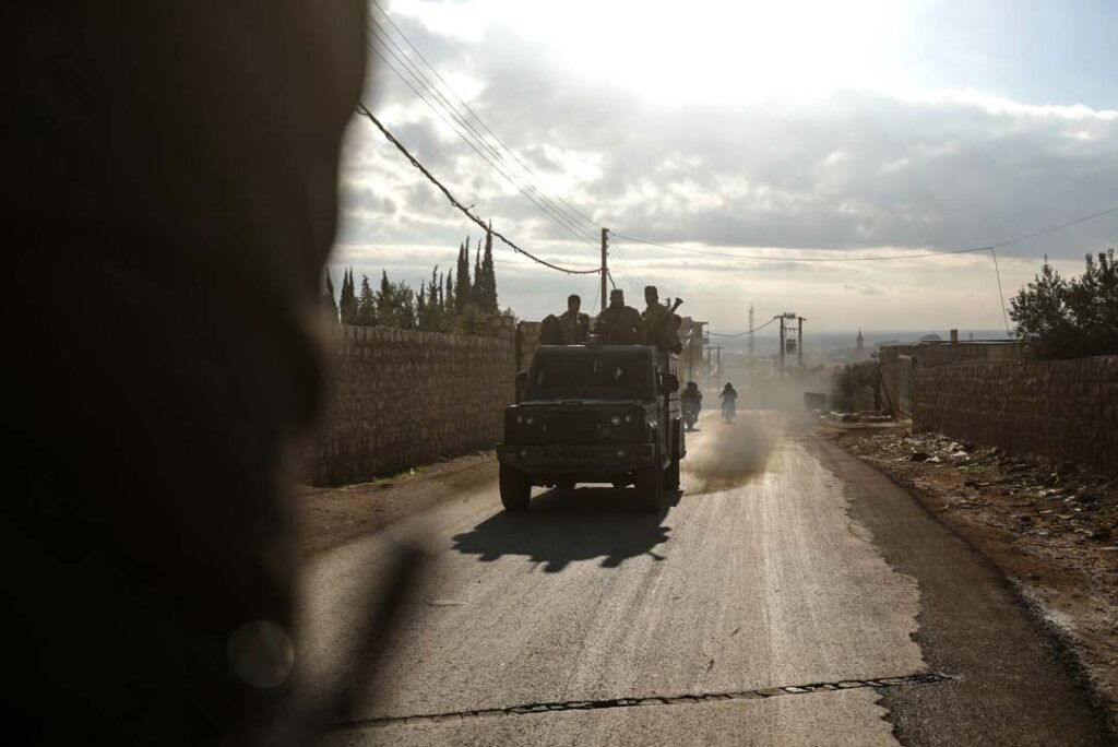 The Struggle to Pacify Syria’s Foreign Legions A military vehicle carrying armed personnel drives down a dusty rural asphalt road flanked by stone walls under a hazy sun, providing visual context to the ongoing conflict dynamics involving Syria's Foreign Legions.