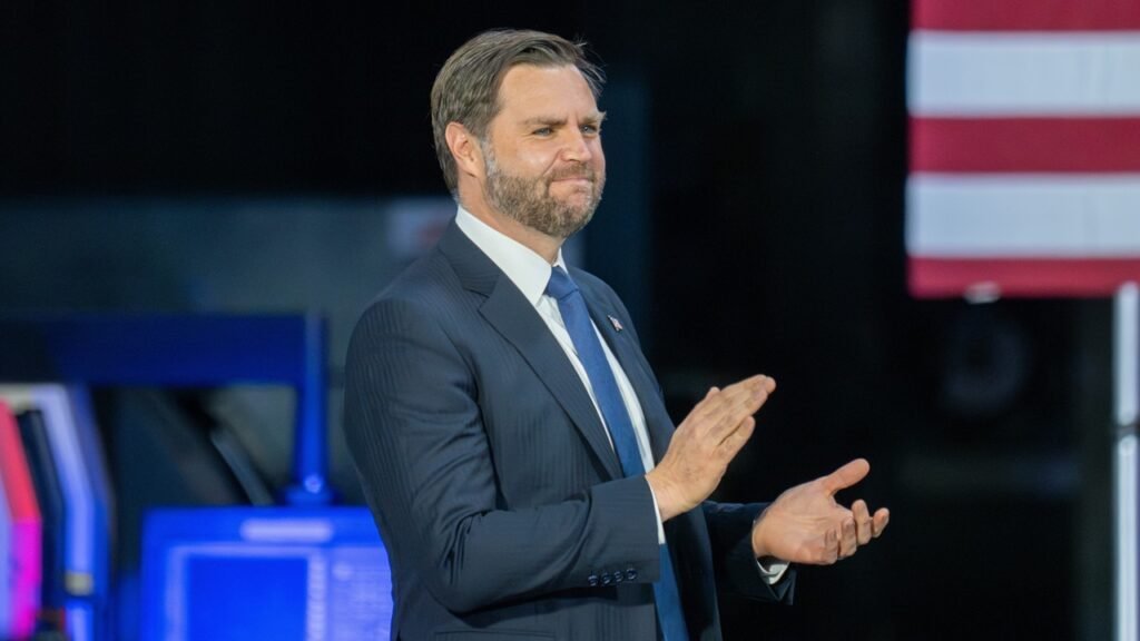 JD Vance Is Wrong: Israel Is Not Just Another Ally JD Vance stands in a dark suit and blue tie, gesturing with his hands during a public event with a portion of the American flag visible in the upper right background, framing the debate around why JD Vance Is Wrong.