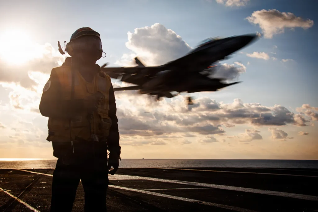 مفارقة القوة في الشرق الأوسط A U.S. Navy crew member stands in silhouette on the flight deck of an aircraft carrier as a fighter jet flies overhead against a cloudy sky at sunset, illustrating the military dynamics involved in the Middle East Power Paradox.