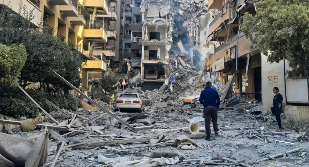 A street scene filled with heavy rubble and debris from a destroyed building surrounded by residential structures and onlookers, capturing the severe infrastructure devastation affecting communities across the Middle East.