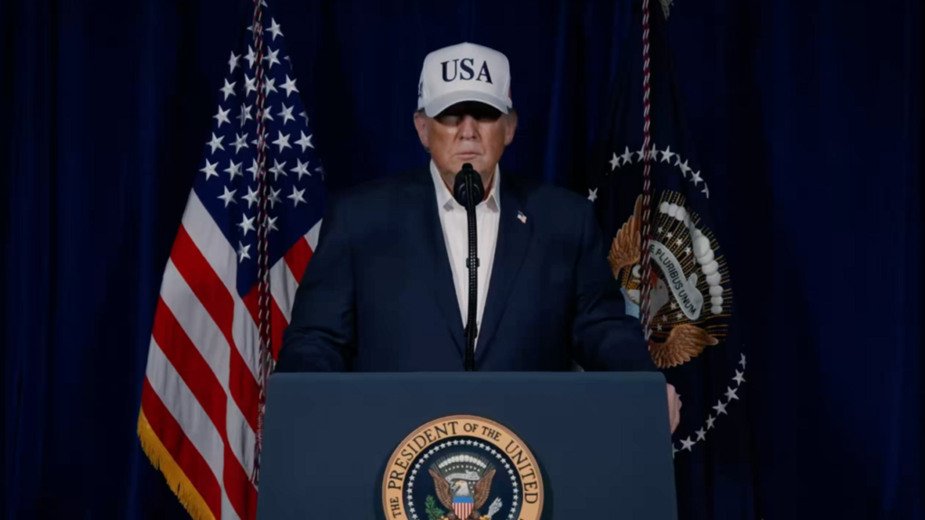 Why No One Trusts the Iran Ceasefire Donald Trump wearing a white USA cap speaks from a podium displaying the presidential seal, flanked by the American flag against a dark blue curtain, providing context to the political backdrop of the finalized Iran ceasefire.