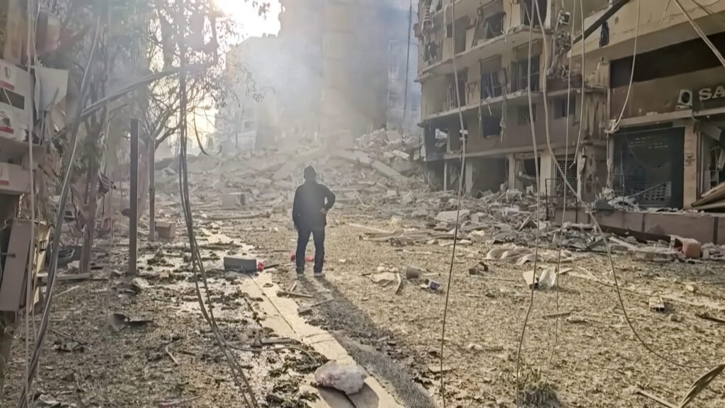 The Middle East’s Regional War Flows Through Lebanon A lone individual stands amidst the dust and heavy rubble of a completely devastated urban street flanked by shattered residential buildings, illustrating the stark human and structural toll of a regional war unfolding in the Middle East.