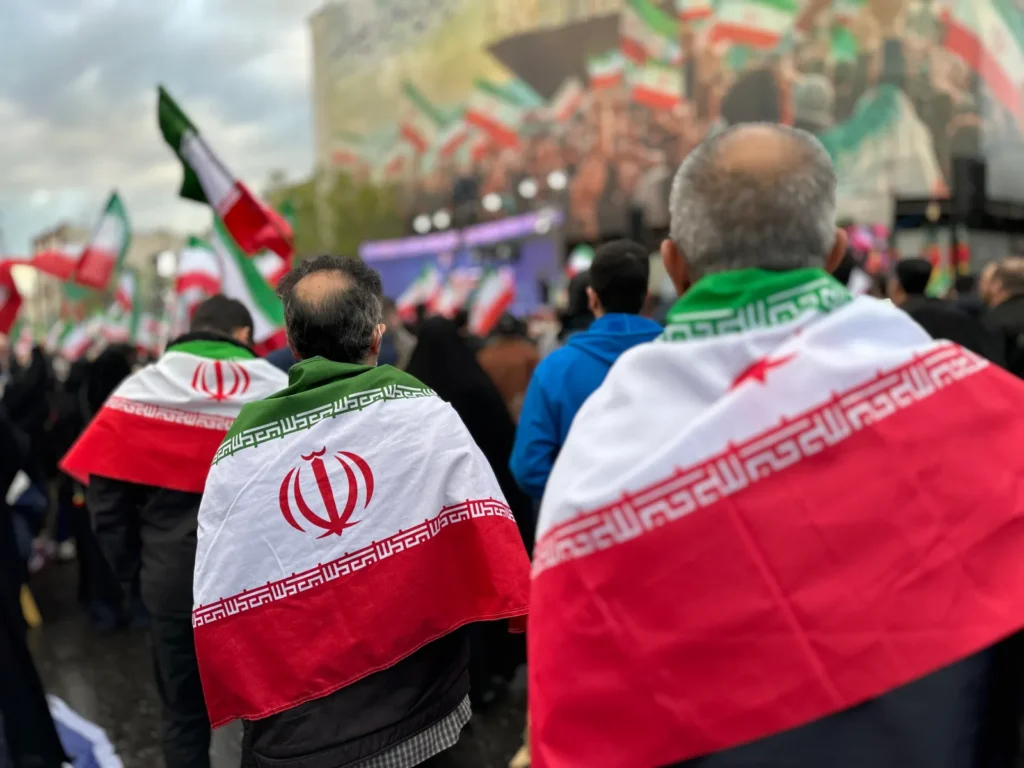 Trump Said US Intelligence Beat Iran—The Record Backs Him A street level view focusing on the backs of demonstrators draped in Iranian flags walking through an outdoor gathering in Tehran, providing visual context to discussions surrounding regional military confrontations and the strategic insights gathered by US intelligence.