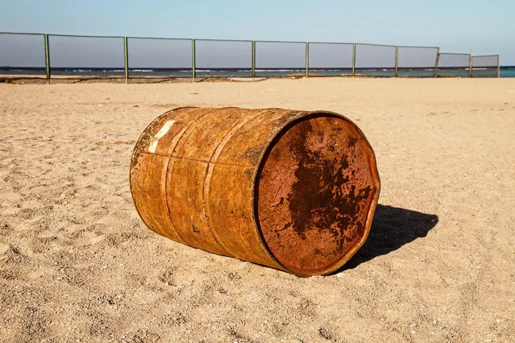 منحدر إمدادات الطاقة يقترب بشكل مقلق A heavily rusted metal oil barrel lies discarded on its side across a vast, empty sandy beach in front of a wire fence and the ocean, visually capturing the stark depletion implied by the looming energy supply cliff.