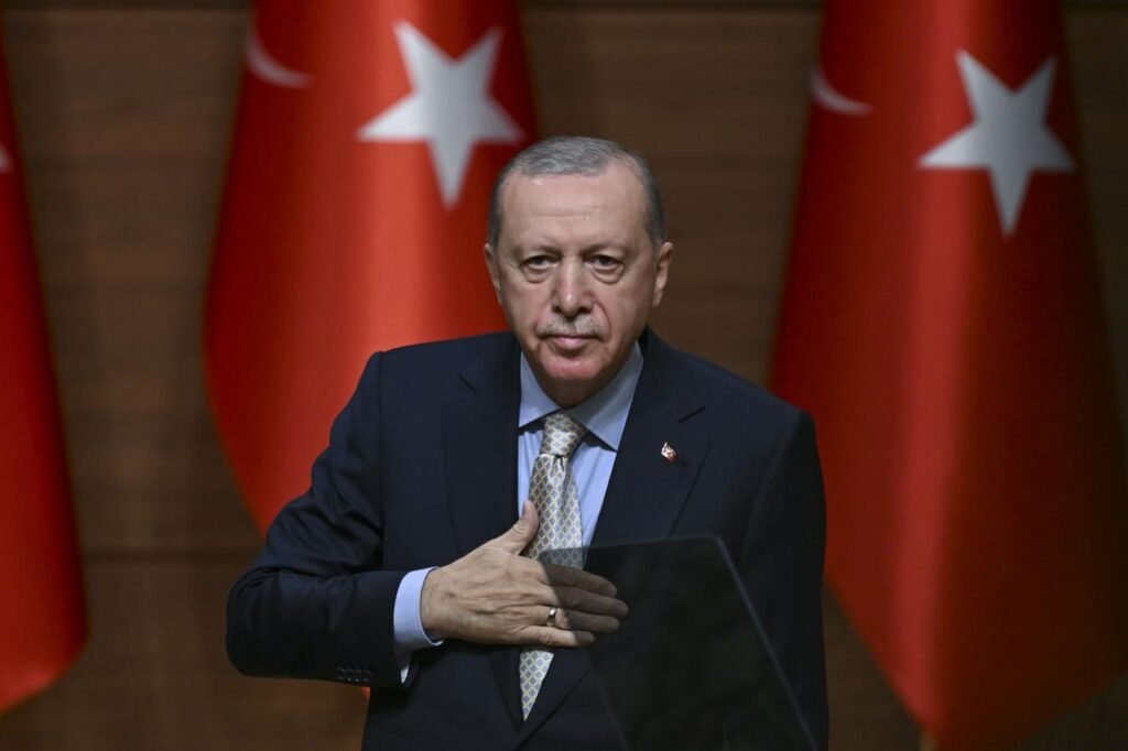 Turkey’s democracy is dying as Erdogan dismantles opposition Turkish President Recep Tayyip Erdogan in a dark suit placing his hand over his heart while standing at a podium, flanked by the national flags of Turkey in the background, illustrating the institutional consolidation affecting Turkey's democracy.