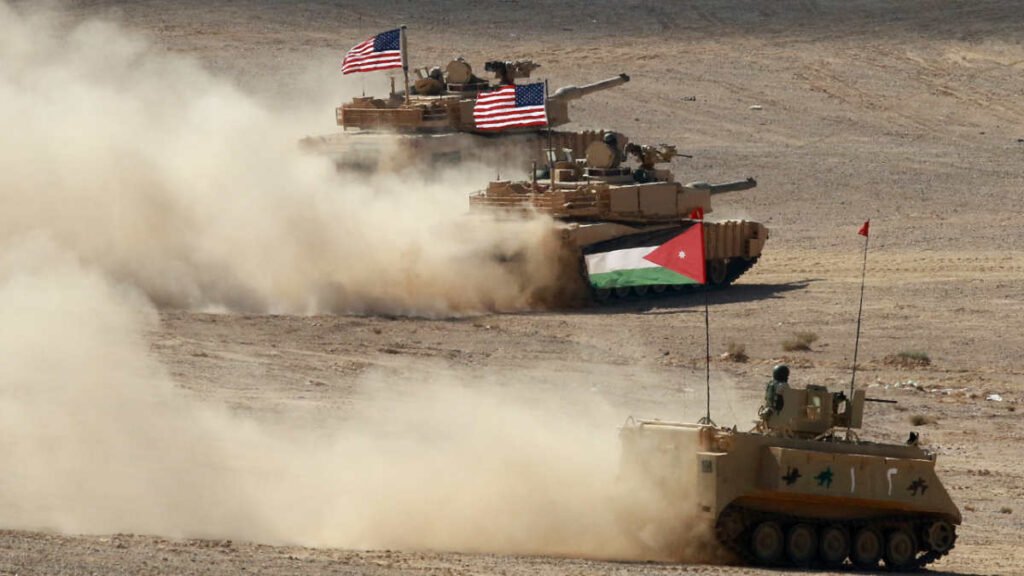ایران خواهان مجازات اردن به خاطر میزبانی از نیروهای آمریکایی است Two American main battle tanks kick up thick desert dust alongside a military armored personnel carrier flying a Jordanian flag, visually illustrating the strategic geopolitical positioning that leads Tehran to threaten to punish Jordan.