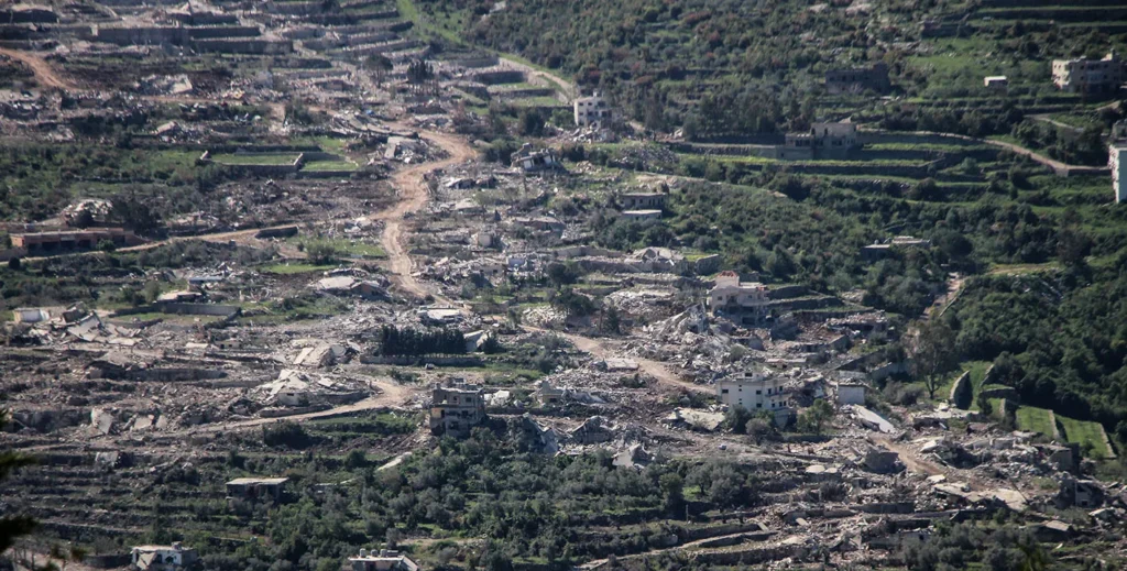 An aerial panoramic view of a heavily damaged rural village with destroyed buildings and debris scattered across terraced hillsides, visually representing the regional devastation underlying discussions on Israel's security.