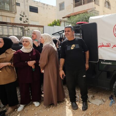 Rebuilding West Bank Refugee Camps: A Shared Interest for Palestinians and Israelis A crowd of residents, including several women wearing headscarves, gathered outdoors near residential buildings and a white emergency utility vehicle with Arabic script, capturing the community environment surrounding the logistical efforts of Rebuilding West Bank Refugee Camps.