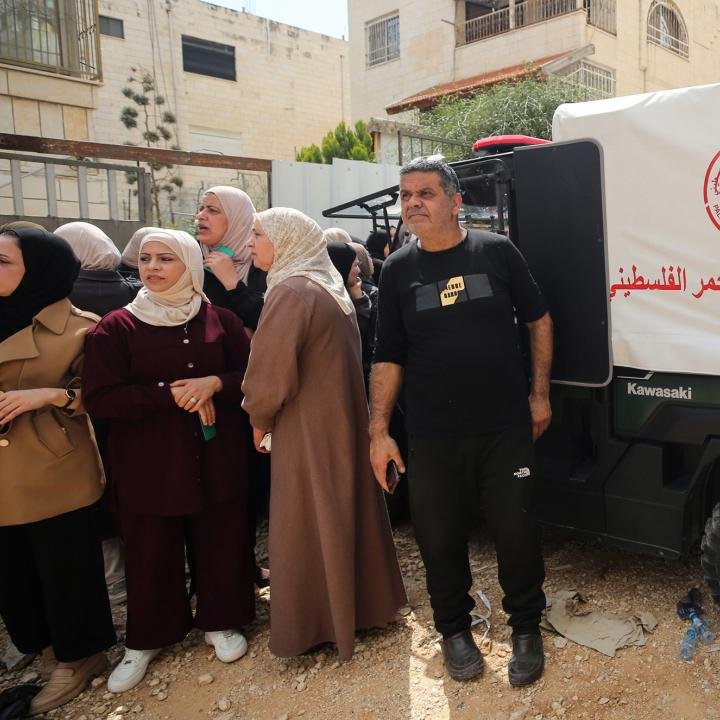 Rebuilding West Bank Refugee Camps: A Shared Interest for Palestinians and Israelis A crowd of residents, including several women wearing headscarves, gathered outdoors near residential buildings and a white emergency utility vehicle with Arabic script, capturing the community environment surrounding the logistical efforts of Rebuilding West Bank Refugee Camps.