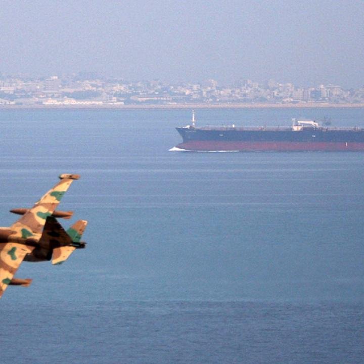 مواجهة أحدث مخطط السيطرة الذكية لإيران في مضيق هرمز A large commercial oil tanker navigates open waters with a coastal city in the distance while the tail wing of a camouflaged military fighter jet appears in the foreground, highlighting the strategic maritime surveillance and security measures associated with Iran's Smart Control in the Strait of Hormuz.