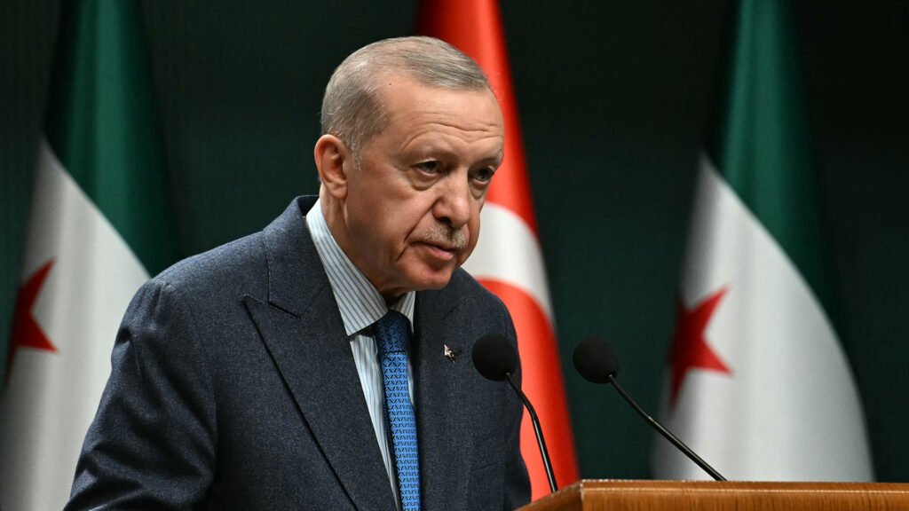 بحث تركيا عن نظام في الشرق الأوسط Turkish President Recep Tayyip Erdogan speaks at a wooden podium with microphones, flanked by the Turkish flag and Syrian opposition flags, highlighting Ankara's diplomatic maneuvers regarding a new Middle East order.
