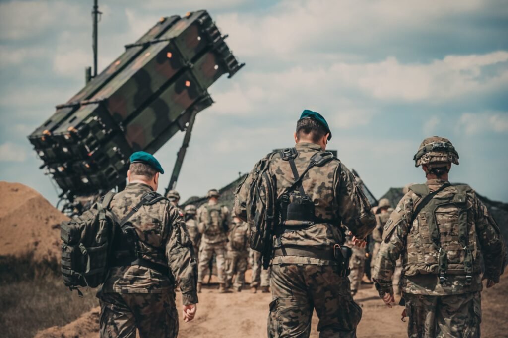 Several soldiers in military camouflage gear walk along a dirt path away from the camera toward a large, angled mobile missile defense battery platform outdoors, illustrating the modern defense infrastructure challenges behind Scaling Patriot Production.