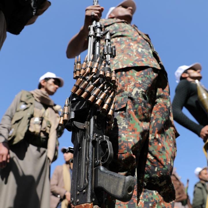 How the Looming Houthi Threat Could Turn the Iran War A low-angle close-up showcases a heavy automatic machine gun wrapped with ammunition belts, held vertically by an armed combatant in camouflage among other forces outdoors, visualizing the expanding regional operational capacity and the Houthi threat.