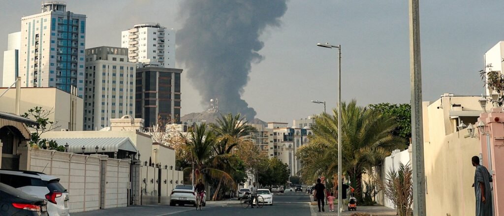 A city street scene in an Arab Gulf state showing modern high-rise buildings, palm trees, and residential walls with a massive column of black smoke rising in the distance, capturing the regional security anxieties surrounding the emerging Iran deal.
