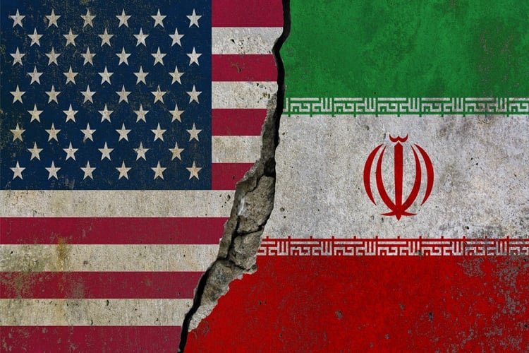 Who Is Responsible for the Iran Debacle? A textured concrete wall painted with the United States flag on the left and the Iranian flag on the right, separated down the middle by a deep jagged crack, metaphorically representing the geopolitical rift and escalation leading to the Iran Debacle.