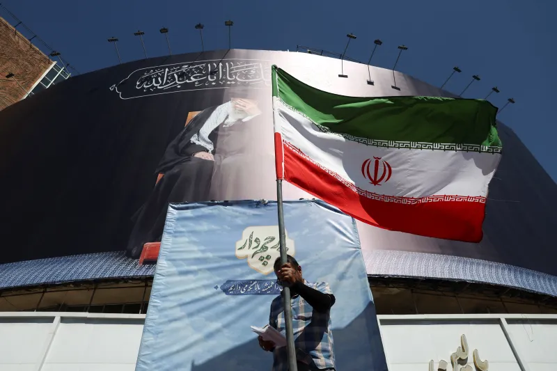 سایه بلند جنگ ایران A man stands holding a large Iranian flag waving in front of massive public billboards in Tehran, capturing the domestic atmosphere defined by The Long Shadow of the Iran War.