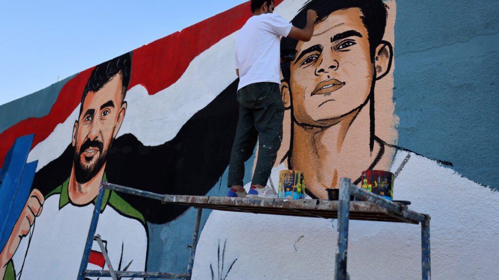 أربعون عاماً في البرية: الطريق الطويل للعراق نحو كأس العالم An artist stands on scaffolding painting a large outdoor street mural featuring portraits of national football players against a stylized Iraqi flag, visually celebrating the athletic milestone of Iraq's World Cup Return.