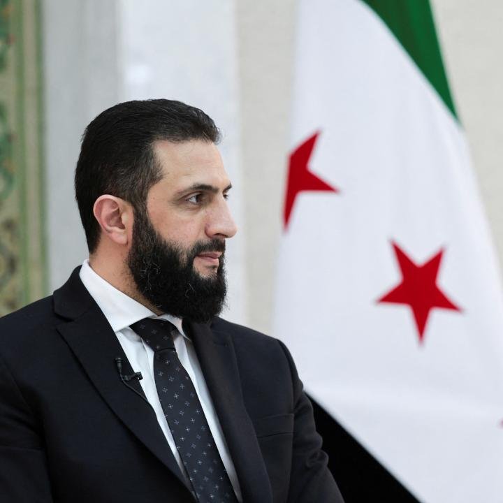 The U.S. Should Not Encourage Syria to Enter Lebanon A profile portrait of Syrian President Ahmed al-Sharaa looking to the side in a black suit and patterned tie, with the Syrian opposition flag blurred in the background, framing discussions on why The U.S. Should Not Encourage Syria.